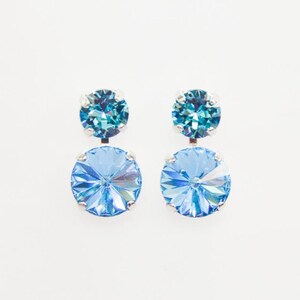 May include: A pair of light blue and teal crystal earrings. Each earring features a small teal crystal on top and a larger, round light blue crystal below. The crystals are set in a silver-colored metal.