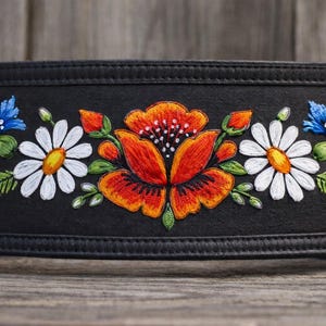 Puede incluir: Cinturón negro con diseño floral bordado. El bordado presenta una flor naranja central con margaritas blancas y amarillas y flores azules a cada lado. El cinturón está hecho de un material oscuro y tiene un borde cosido.