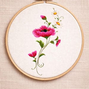 Puede incluir: Arte floral bordado en bastidor con una amapola rosa vibrante y delicadas margaritas blancas. Las flores están sobre un fondo de tela blanca dentro de un bastidor de bordado de madera. Hojas y tallos verdes completan el diseño.