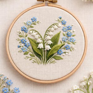 Puede incluir: Arte floral bordado en un aro de madera. Presenta un grupo central de flores blancas de lirios de los valles con hojas verdes, rodeado de nomeolvides azules. La bordado está en un aro de madera.