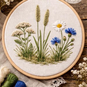 Può includere: Ricamo floreale su telaio con una varietà di fiori selvatici, tra cui margherite e fiordalisi, nei toni del bianco, blu e verde. Il testo "PDF PATTERN" è visibile. Il ricamo è posto in un telaio di legno.