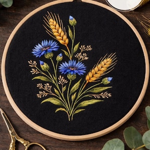 May include: An embroidery hoop with a black fabric featuring a floral design of blue cornflowers and golden wheat. The text "PDF pattern" is in a white circle. Gold scissors are in the lower left corner.