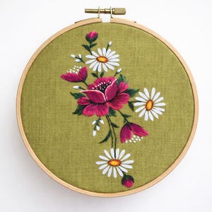 Puede incluir: Diseño floral bordado en un aro de madera. El diseño presenta una flor grande, de color rosa intenso, con detalles blancos, rodeada de margaritas blancas y capullos rosas más pequeños, todo sobre un fondo de tela verde. El aro de madera se asegura con un cierre de latón.
