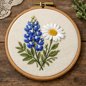 Puede incluir: Arte de aro floral bordado con un altramuz azul y una margarita. El altramuz tiene pétalos azules y blancos, mientras que la margarita tiene pétalos blancos y un centro amarillo. El bordado está en un aro de madera.