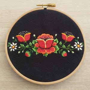 Puede incluir: Un bastidor de bordado redondo de tela negra con un diseño floral. El bordado presenta flores rojas y naranjas, hojas verdes y margaritas blancas. El bastidor de madera tiene un tornillo de ajuste de latón.