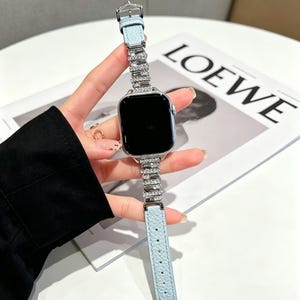 May include: A light blue leather watch band with a silver watch face and a sparkling rhinestone band. The watch band is held by a person. The watch face is black.