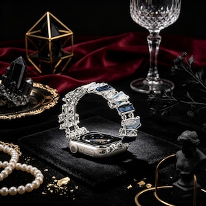 Op de afbeelding: Een zilveren Apple Watch met een decoratieve band met blauwe en heldere accenten. De band is gemaakt van metaal en heeft een geometrisch ontwerp. Het horloge wordt gepresenteerd op een zwart fluwelen oppervlak, met sieraden en decoratieve items op de achtergrond.