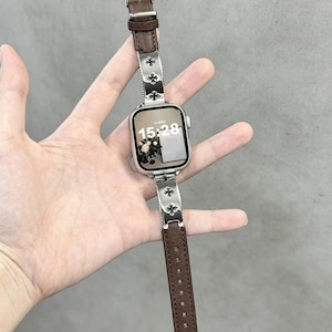 以下が含まれることがあります： 茶色の革バンドが付いたシルバーのスマートウォッチ。バンドには、花の模様の装飾的なシルバーのアクセントが付いています。時計の文字盤には15:28と表示されています。背景はニュートラルです。