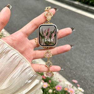 May include: Gold Apple Watch with a decorative strap. The strap has pink floral accents and clover shapes. The watch face shows a floral image and the time 15:48. The watch is a stylish accessory.