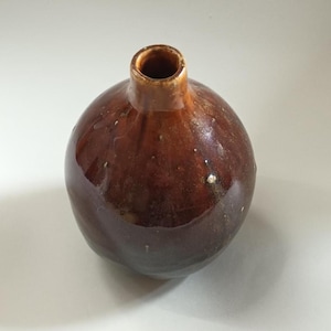 Può includere: Un piccolo vaso in ceramica rotondo con una smaltatura lucida color marrone scuro. Il recipiente presenta un collo stretto e cilindrico e un corpo bulboso con sottili rientranze organiche sulla superficie, che riflettono la luce.