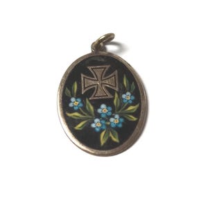 May include: Oval pendant with a silver-colored frame and a black enamel background. The pendant features a silver cross at the center, surrounded by blue forget-me-not flowers and green leaves. The pendant has a small loop at the top.