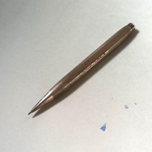 Può includere: Una matita meccanica argentata con un corpo strutturato a motivo di diamanti. La matita ha una punta affilata e una piccola iscrizione in corsivo. La matita è appoggiata su una superficie bianca.