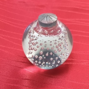 Puede incluir: Un pisapapeles de vidrio transparente con forma de esfera y parte superior plana. El interior del vidrio contiene numerosas burbujas de aire pequeñas y suspendidas dispuestas en un patrón. La parte superior tiene una tapa gris texturizada.