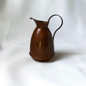 Op de afbeelding: Een kleine, koperkleurige kan met een gebogen handvat. De kan heeft een afgeronde body en een tuit om uit te schenken. Het handvat is een donkerdere bruintint. De kan is waarschijnlijk vintage en geschikt voor decoratieve doeleinden.