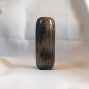 Könnte beinhalten: Ein zylindrischer, brauner Flaschenverschluss mit glänzendem Finish. Der Verschluss hat eine abgerundete Oberseite und ein dunkles, gesprenkeltes Muster. Die Farbe geht von hellbraun zu dunkelbraun und schwarz über. Es ist ein dekorativer Artikel.