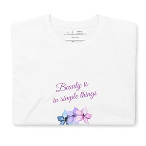 Può includere: Una t-shirt bianca con il testo Beauty is in simple things stampato in un carattere corsivo viola. Sotto il testo ci sono tre fiori illustrati nelle tonalità del blu, viola chiaro e rosa. La maglietta presenta un'etichetta con taglia S.