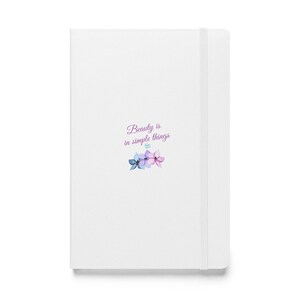 Può includere: Un taccuino con copertina rigida bianca e fascia elastica di chiusura. La copertina presenta la scritta Beauty is in simple things in corsivo viola, accompagnata da una piccola illustrazione di tre fiori in stile acquerello blu, viola e rosa.