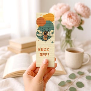 Pode incluir: Marcador de livros bege com um desenho de ponto cruz de uma abelha e o texto "BUZZ OFF!". A abelha é verde-azulado, laranja e castanha, com círculos laranja e amarelos por cima. Fundo desfocado com livro, flores e caneca.