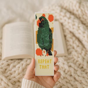 May include: A handmade bookmark featuring a pixelated parrot design in shades of green, orange, and black. The bookmark has the text "REPEAT THAT" in yellow. The bookmark is held in front of an open book.