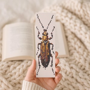 May include: A handmade bookmark featuring a detailed cross-stitch design of a beetle in shades of brown, gold, and black. The bookmark is rectangular with a white background and is held in front of an open book and a cream-colored blanket.