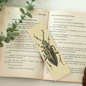 May include: Beige cross-stitch bookmark featuring a detailed green and yellow beetle design. The bookmark is placed in an open book with text from Jules Verne's "Around the World in Eighty Days".