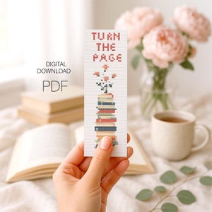 May include: A white bookmark with a cross-stitch design. The bookmark shows a stack of books with flowers and the text "TURN THE PAGE" in red. The words "DIGITAL DOWNLOAD PDF" are also visible. The bookmark is held in a hand.