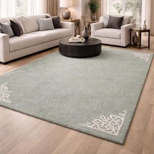 May include: A large, rectangular, light green rug with a soft texture. The rug features a white, ornate corner design. The rug is placed on a light wood floor in a living room setting.