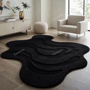 Puede incluir: Una alfombra grande, negra, de forma abstracta con un diseño texturizado en forma de onda. La alfombra está colocada sobre un suelo de madera clara en un salón moderno. La forma única y el color oscuro de la alfombra crean un contraste llamativo.