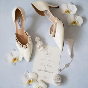 Peut inclure: Escarpins Jimmy Choo ivoire avec perles, invitation de mariage, bagues et orchidées blanches sur fond bleu clair. Une clé décorative et un petit buste complètent le thème nuptial.