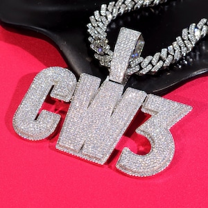 May include: A diamond-encrusted pendant spelling out "GW3" with a matching chain. The pendant and chain are silver-colored and set against a red background. The jewelry is highly reflective, catching the light.
