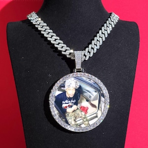 May include: A silver-toned chain necklace with a large, circular pendant. The pendant has a photograph of a person inside a car, encircled by small, clear stones. The chain is made of interlocking links, and the necklace is displayed on a black stand.