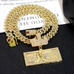 May include: A gold-colored, diamond-studded chain necklace with a matching pendant. The pendant is shaped like a dollar bill with a portrait and the word "MOMENT" on the top. The chain is made of interlocking links.