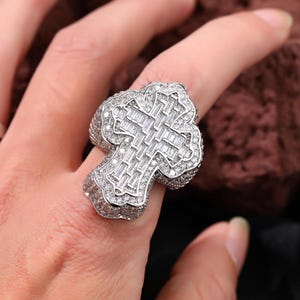 May include: A silver-colored ring in the shape of a cross, encrusted with numerous small, clear gemstones. The cross design features a central section of rectangular stones. The ring is worn on a finger.