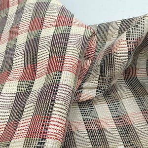 Puede incluir: Primer plano de una tela tejida con un patrón de cuadros. La tela presenta una mezcla de colores, incluyendo rojo, negro, crema y marrón, dispuestos en rayas verticales y horizontales. La tela está doblada, mostrando su textura y caída.