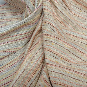 Puede incluir: Primer plano de una tela beige con un patrón de rayas. La tela tiene finas rayas horizontales en tonos rojos, naranjas, azules y amarillos. La tela está drapeada, mostrando pliegues y textura, lo que sugiere que es un material tejido.