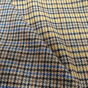 Op de afbeelding: Een geweven stof met een meerkleurig glen plaid patroon. Het textiel vertoont een herhalend ruitontwerp bestaande uit kruisende lijnen in tinten mosterdgeel, bruin, zwart en felblauw, wat zorgt voor een getextureerd uiterlijk van de stof.