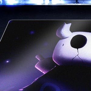 May include: A rectangular desk mat featuring a dark purple and black gradient background with a stylized white character head. The character has large circular black eyes, a cracked surface texture, and small horn-like protrusions on the top.