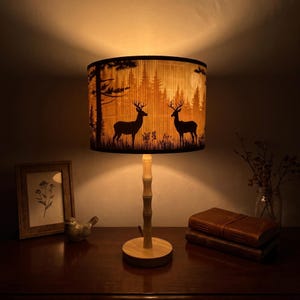 Può includere: Una lampada da tavolo con paralume cilindrico che presenta la sagoma nera di due cervi in un bosco. La lampada ha uno stelo color chiaro in stile bambù e una base circolare in legno, che emette una luce calda e soffusa dal paralume.