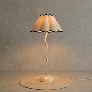 May include: A cream-coloured table lamp with a wavy stem and a scalloped lampshade. The lampshade is cream-coloured with a black trim. The lamp sits on a round, woven mat. The lamp is plugged into an electrical outlet.