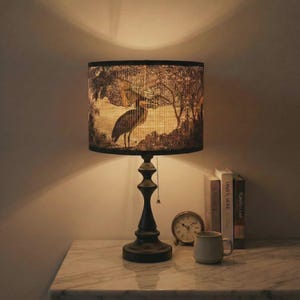 May include: A table lamp with a black base and a lampshade depicting a bird in a natural setting. The lamp sits on a marble-effect surface, accompanied by books, a small clock, and a mug. The lamp has a pull chain.