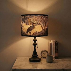 May include: A black table lamp with a round lampshade featuring a heron illustration. The lamp has a dark base and a pull chain. A small clock, books, and a mug are on the table next to the lamp.