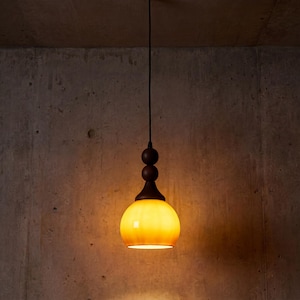 Puede incluir: Una lámpara colgante con una pantalla de globo de cristal amarillo-naranja. La lámpara tiene una parte superior decorativa marrón oscuro con tres acentos esféricos y un cable negro. Cuelga contra una pared de hormigón gris texturizado.