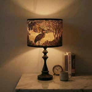 May include: A decorative table lamp with a black base and a lampshade featuring a nature scene with a heron. The lamp is on a marble-topped table, next to books, a small clock, and a mug. The lamp's shade has a black border.