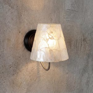 Peut inclure: Applique murale avec abat-jour blanc cassé texturé. L'abat-jour a un aspect superposé, et le luminaire est fixé à une base circulaire foncée. L'applique est fixée sur un mur gris texturé.