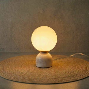 May include: A small, spherical table lamp with a warm, glowing light. The lamp features a round, frosted glass shade atop a beige, rounded base. The lamp is placed on a circular, woven mat.
