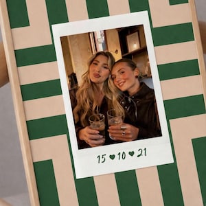 Könnte beinhalten: Ein gerahmter Druck mit einem Foto im Polaroid-Stil von zwei Personen, die Getränke halten. Das Foto ist vor einem grün-beige gestreiften Hintergrund platziert. Das Datum "15 10 21" ist auf dem Foto aufgedruckt.