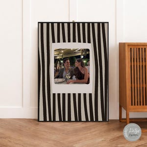 Könnte beinhalten: Ein gerahmtes Foto mit einem schwarz-weiß gestreiften Rand. Das Foto im Inneren zeigt ein Paar in einem Restaurant. Der Rahmen ist rechteckig und hat einen weißen Innenrand. Das Foto ist ein Polaroid-Druck.