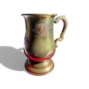 May include: An antique brass mug with a decorative handle and base. The mug features an embossed emblem on the front. The mug is approximately 6 inches tall.