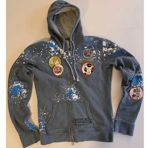 May include: Gray zip-up hoodie with a hood and front pockets. The hoodie features white and blue paint splatter designs and several circular patches with illustrations. The text "DON ED HARDY designs" is printed on the front.