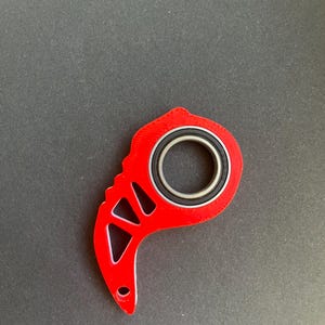 May include: A red, geometric-shaped fidget spinner with a circular bearing in the center. The spinner has a curved handle with a hole at the end and triangular cutouts. The bearing has a black rubber ring and a silver inner ring.